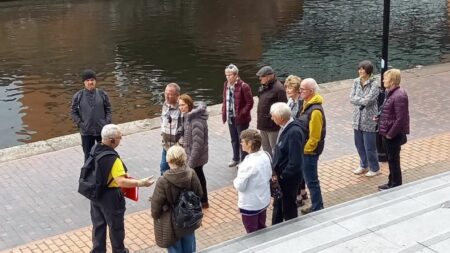 A landscape image showing a group of 12 people on a canal towpath being spoken to by a person in a yellow t shirt. In front of them are some grey steps and behind them is a large body of water. The background is yellow. In the bottom right of the image is a yellow smiley face logo and a red square with the letter h in.