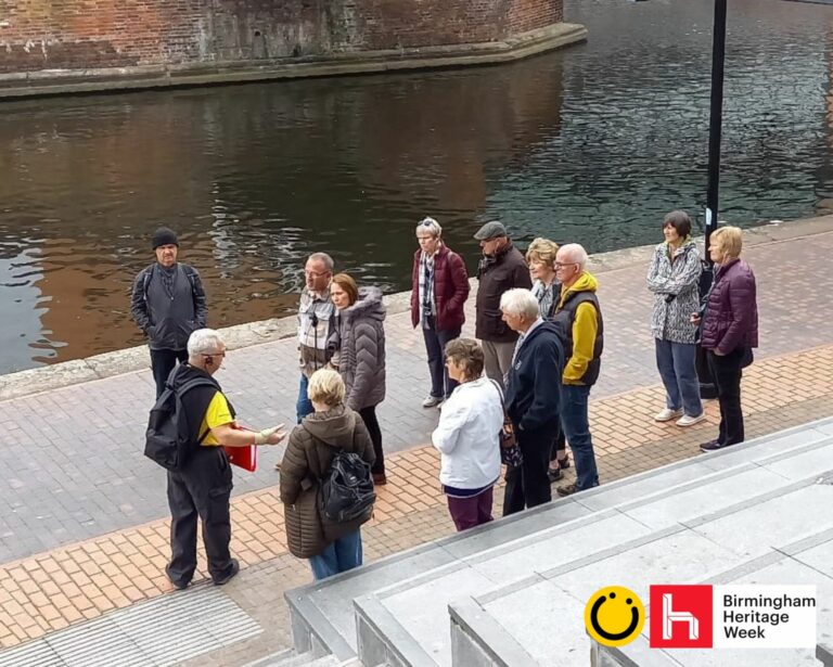 A landscape image showing a group of 12 people on a canal towpath being spoken to by a person in a yellow t shirt. In front of them are some grey steps and behind them is a large body of water. The background is yellow. In the bottom right of the image is a yellow smiley face logo and a red square with the letter h in.
