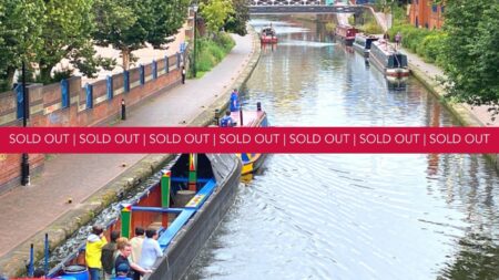 A landscape image showing a view of a canal. Either side of the canal is a path and, on the right, there are people walking along it. On the bottom left part of the image and on the canal is a red roofed and blue and yellow sided canal boat pulling a blue, black, green, red and yellow painted open air boat with 7 people stood on it. In the distance is a black and white metal bridge over the canal and other canal boats on the water. In the bottom right of the image is a yellow smiley face logo and a red square with the letter h in. Across the middle of the image is a red banner with the words sold out on.