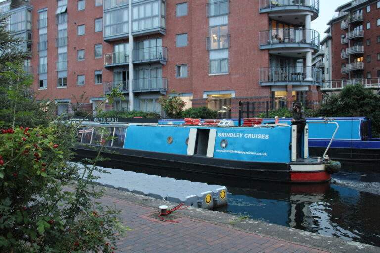 A landscape image showing a blue canal boat on a body of canal water. In the foreground of the image is a path, and on the water next to the path is a grey pontoon.