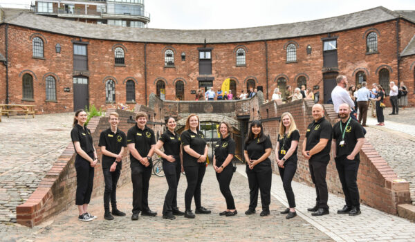 The Roundhouse team in Roundhouse black t-shirts, lined up in the Roundhouse courtyard , with the Roundhouse building and the tunnel in the background