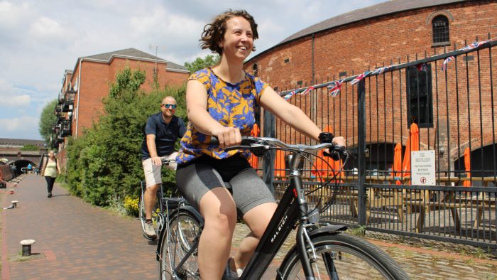 Picture of a lady cycling in the sun, smiling during the Roundhouse cycling. Roundhouse tour guide cycling behind her.